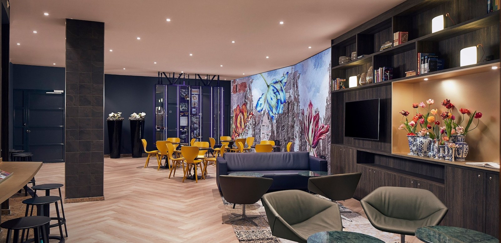 A modern living room with a dark blue wall, a large mural, a flat-screen TV, and a bookshelf with decorative items. - Inntel Hotels Amsterdam Centre (Amsterdam)