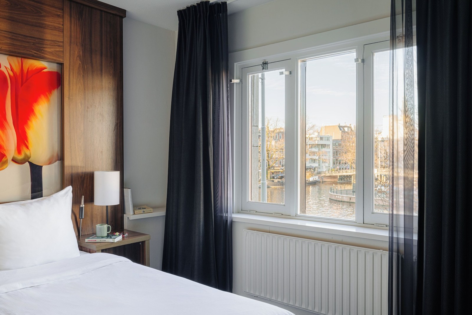 A hotel room with a bed, a nightstand, a lamp, a window with curtains, and a view of buildings and water. - Leonardo Eden Hotel Amsterdam City Center (Amsterdam)