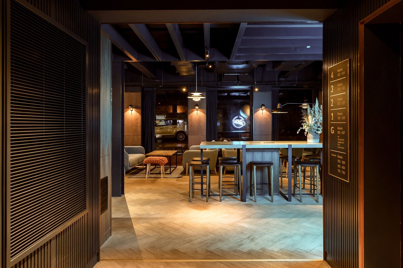 A modern bar area with a wooden bar counter and stools, a seating area with a sofa and ottoman, and a neon sign above the bar. - Wilde Aparthotels, Edinburgh, Grassmarket (Edinburgh)